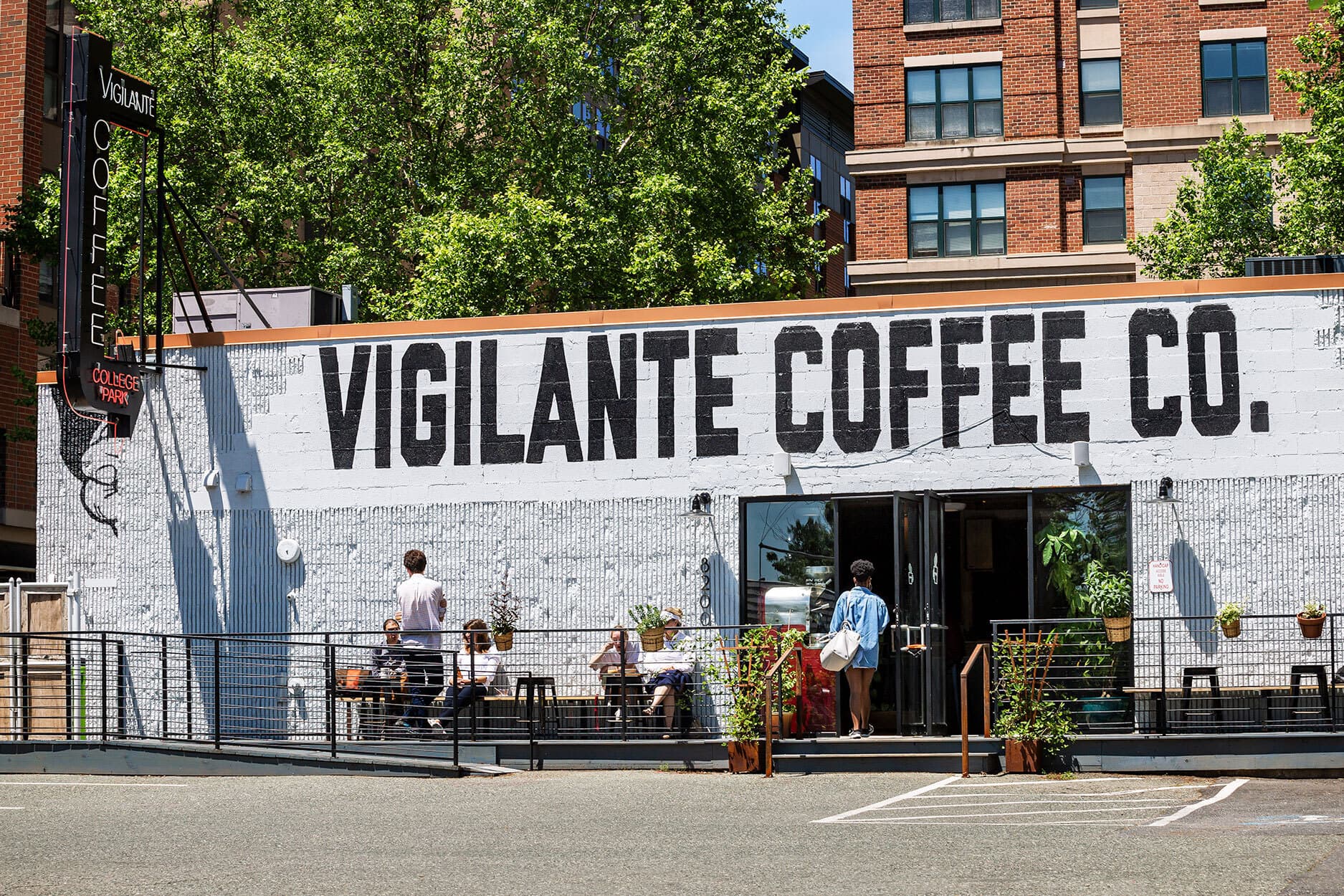 a White-wash store front with plantings by glass frontdoors. The establishment's name in large bolded font stretched across the top of the building: Vigilante Coffee Co.