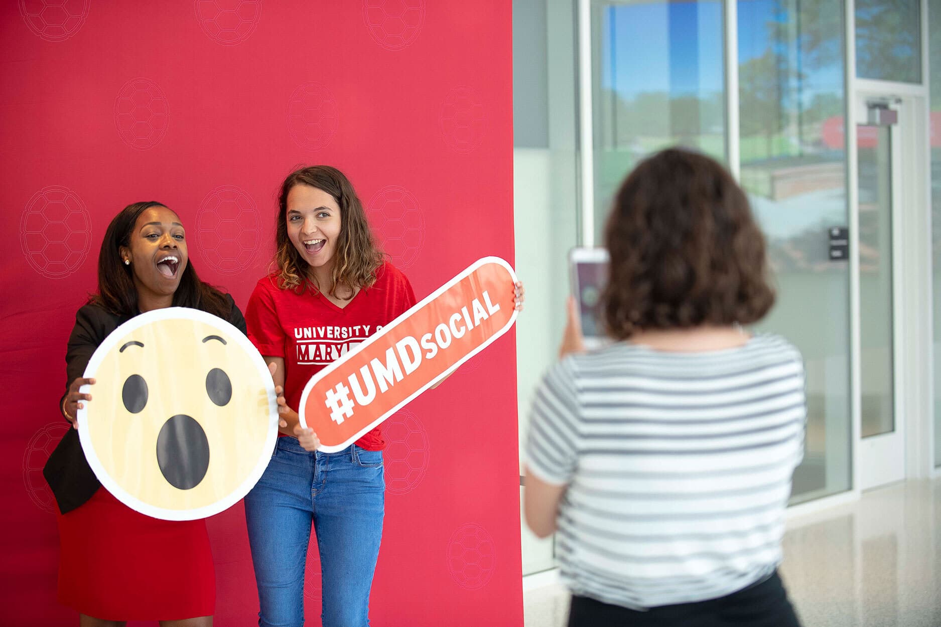 Two conference attendees take a photo while holding props of the Wow face emoji, and the event's hashtag UMDSocial