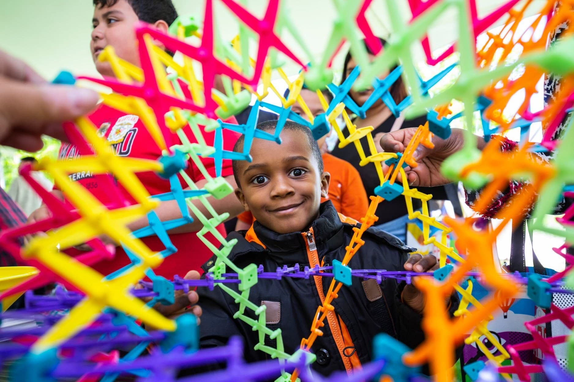A young person sits on the other side of a very colorful expanded Hoberman sphere looking straight at the camera
