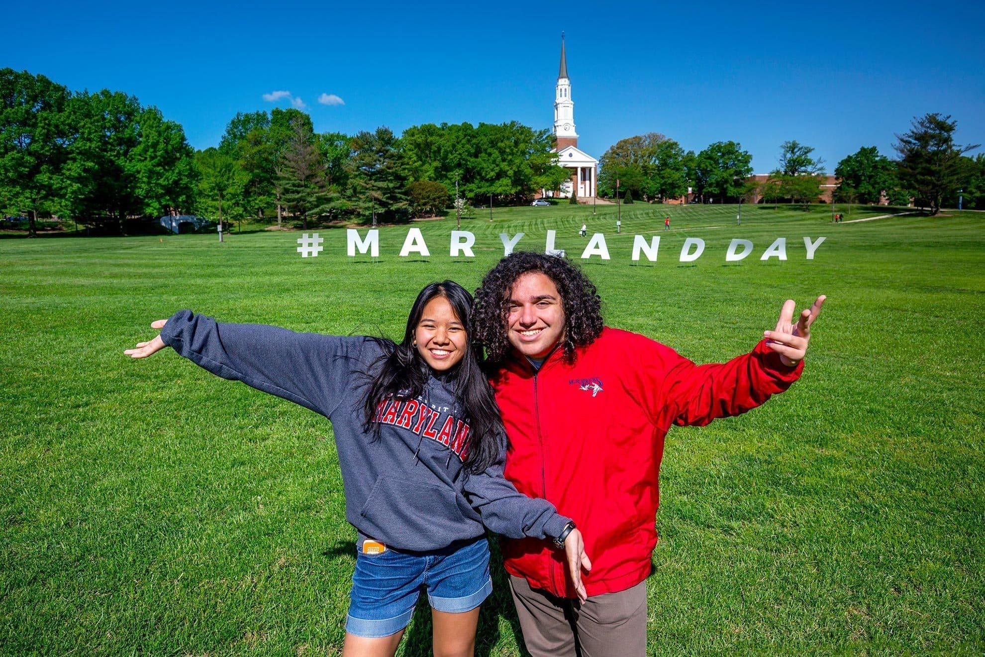 Two Maryland Day attendees stand on Chapel Field showcasing the huge Maryland Day sign
