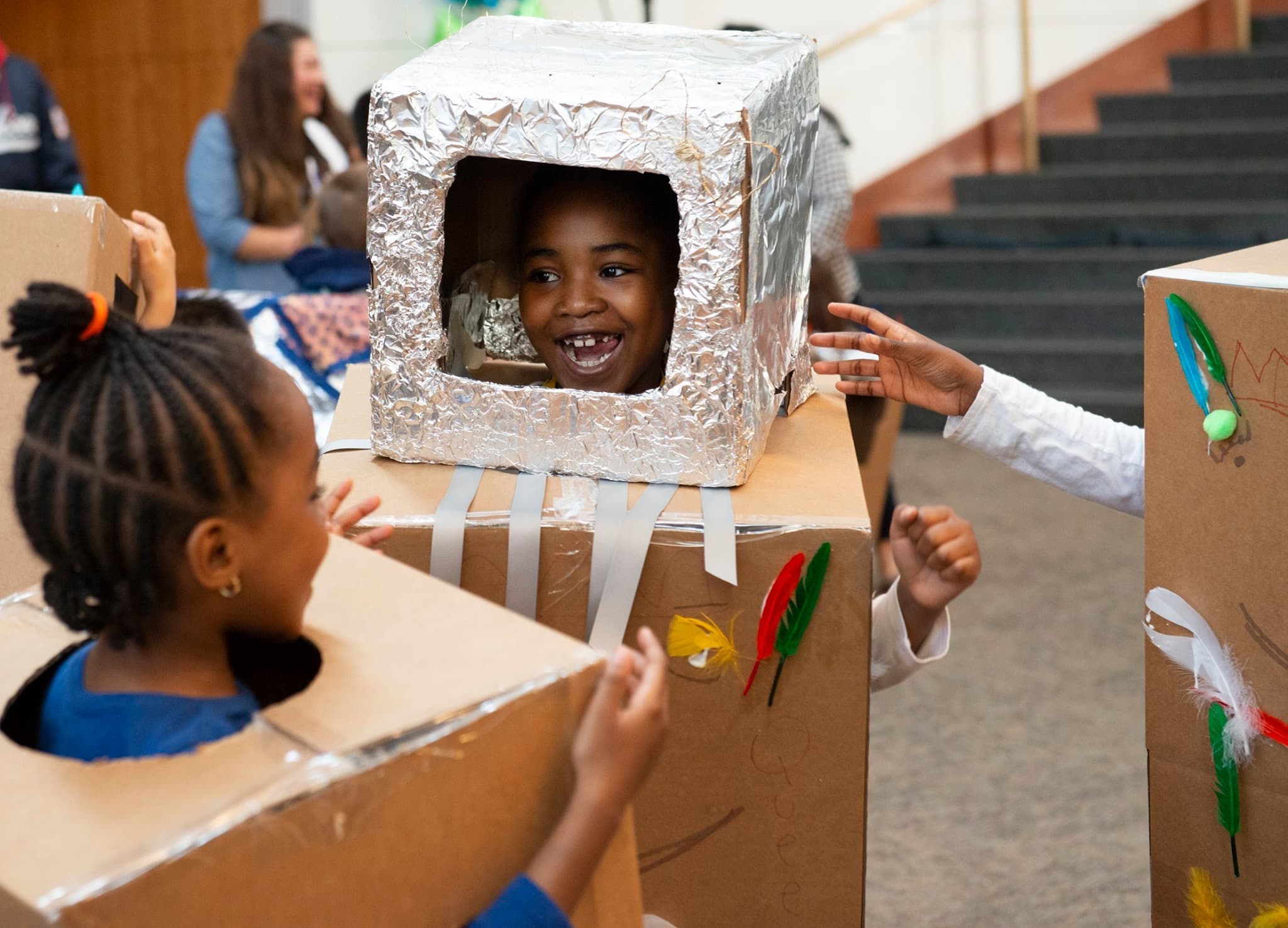 A group of young people smile and laugh together while sporting cardboard boxes around their torsos and head covered in tape tinfoil and various craft supplies as if they are robots