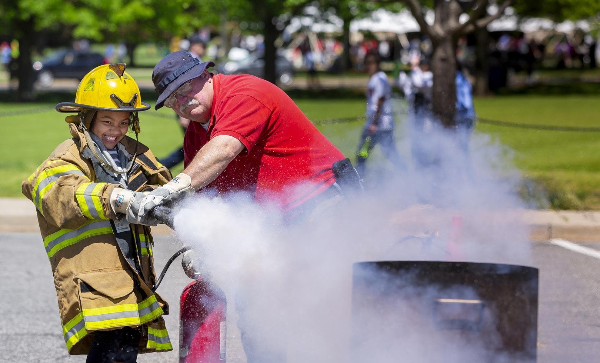 A child wearing an oversized adult fire fighter coat and helmet is helped by an instructor as they spray an open flame with a fire extinguisher