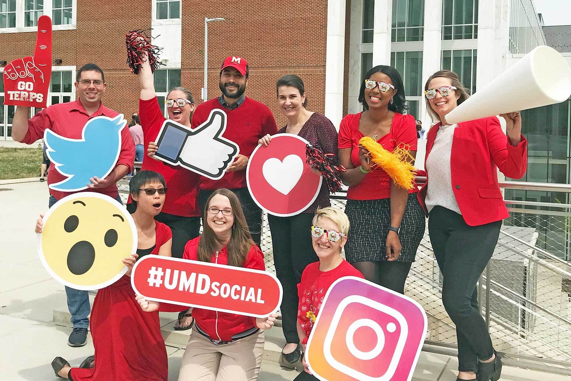 Group photo of #UMDSocial attendees all holding enlarged printed versions of social media related emojis like the facebook heart, twitter logo, and a megaphone graphic.